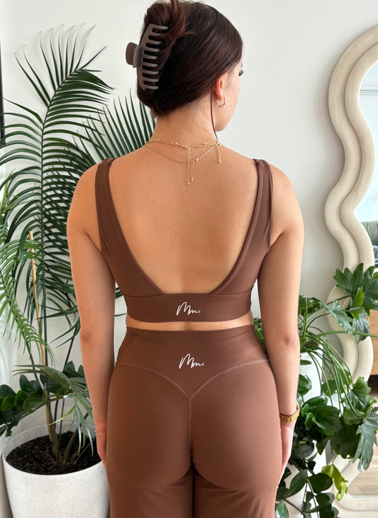 Person wearing a brown sports bra and leggings set with 'Mia' branding, standing in a room with plants.